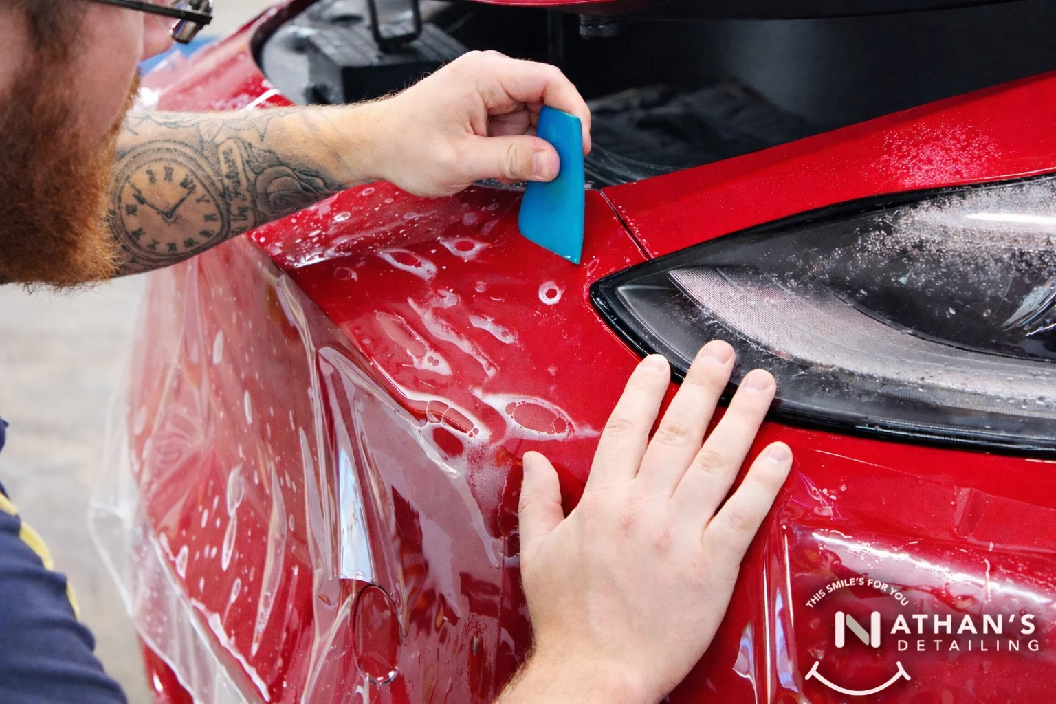 Squeegeeing paint protection film along headlight trim on a red car.