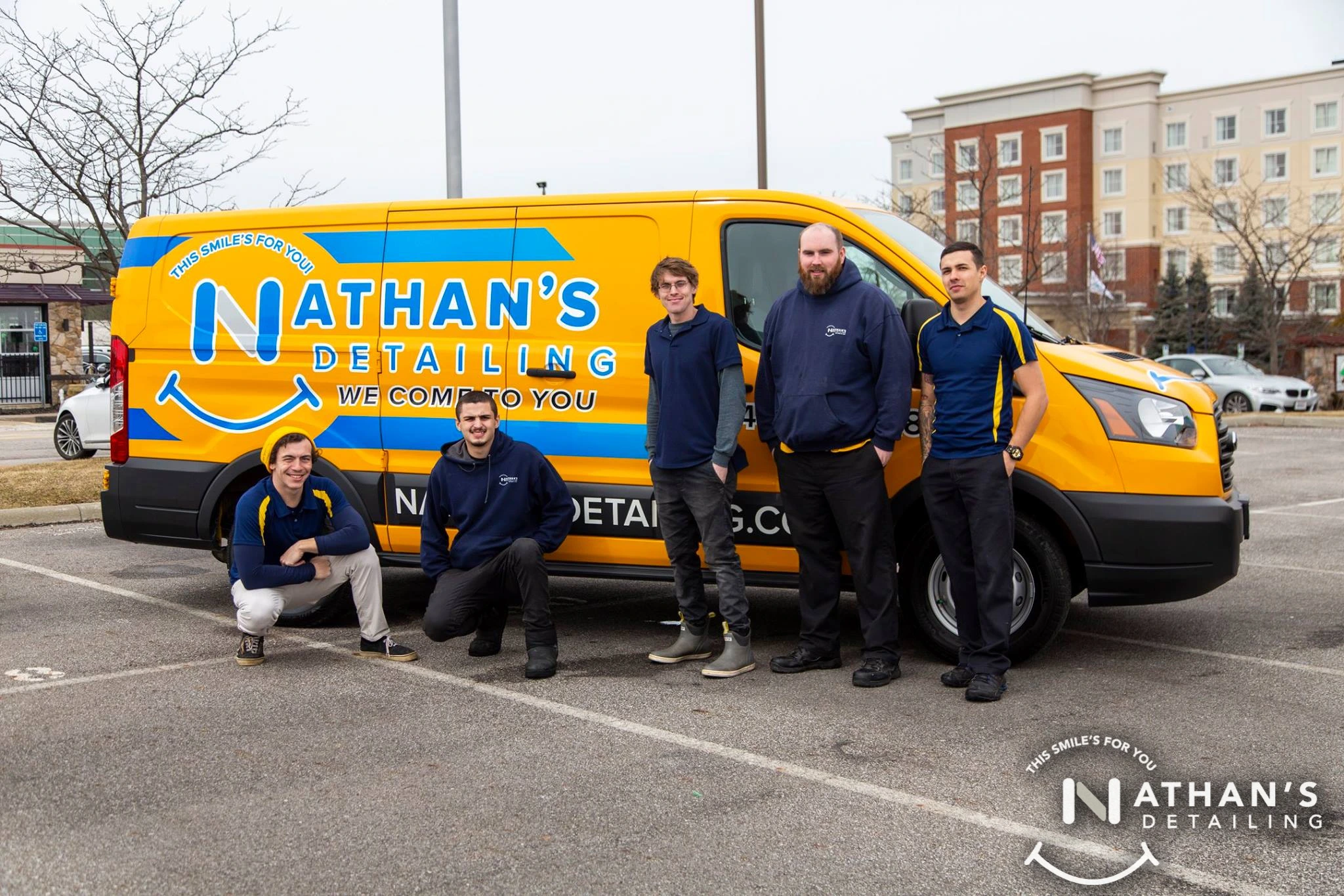 Five men in navy and yellow uniforms pose in front of a yellow Nathans Detailing van in a parking lot, with buildings and trees in the background. The van has the company’s logo and slogan on the side.