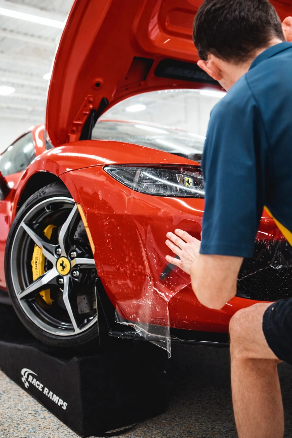 A person applies a clear protective film to the front bumper of a red Ferrari sports car, which is raised on black Race Ramps. The cars hood is open, and the focus is on the vehicles front wheel and headlight.