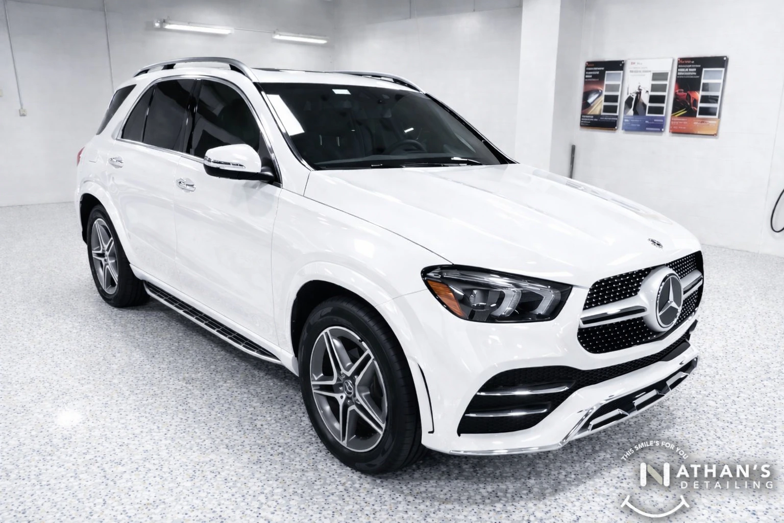 A white Mercedes-Benz SUV is parked in a clean, brightly lit detailing garage with tiled floors. Sample paint colors and detailing products are visible on the wall in the background. The Nathan’s Detailing logo is in the corner.