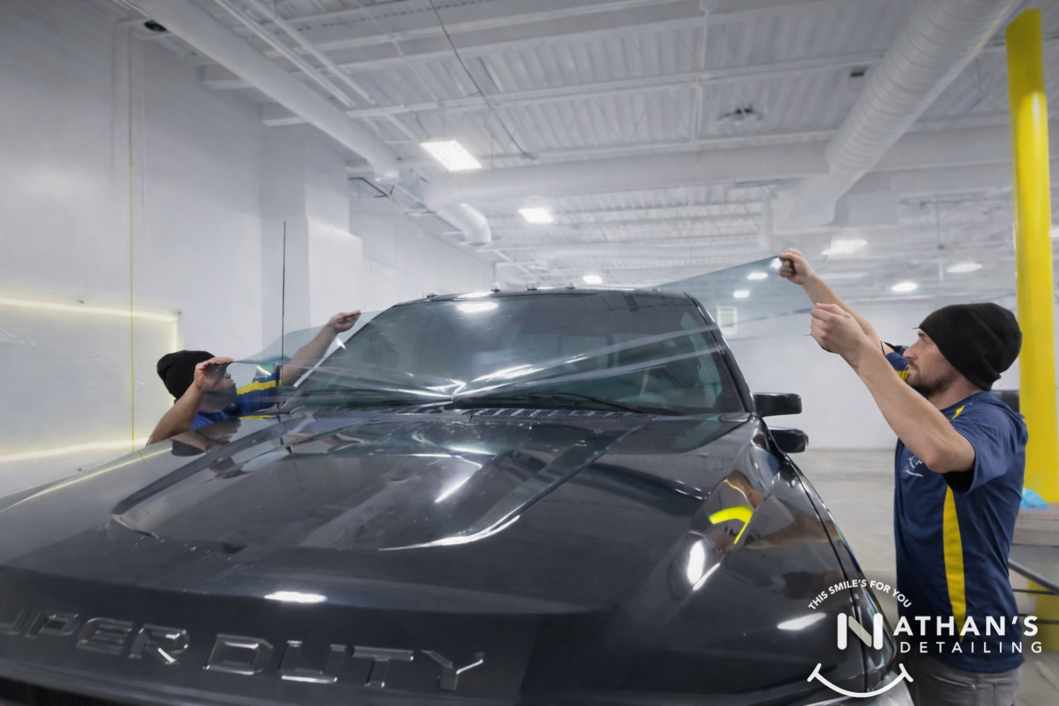 Two technicians stretching tint film across a truck windshield in a clean shop.