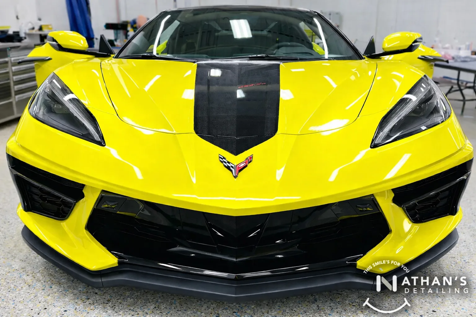 A bright yellow Chevrolet Corvette with a black racing stripe is parked indoors on a speckled garage floor. The cars front view showcases its sleek, sporty design. A Nathan’s Detailing logo appears in the bottom right corner.