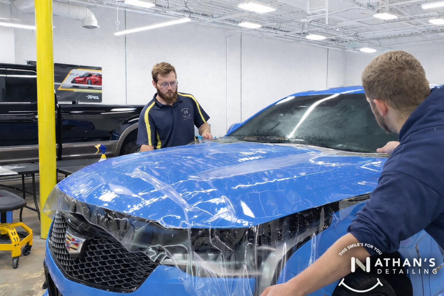 Two installers positioning paint protection film across a blue vehicle hood in Chagrin Falls, Ohio.