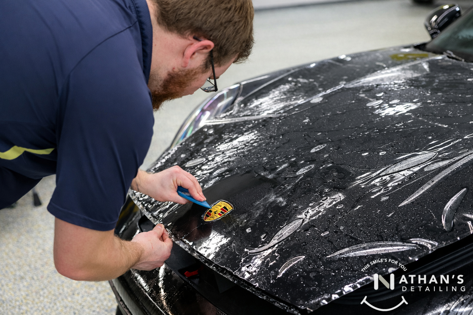 Close-up of paint protection film being trimmed around a Porsche hood badge in Chagrin Falls, Ohio.