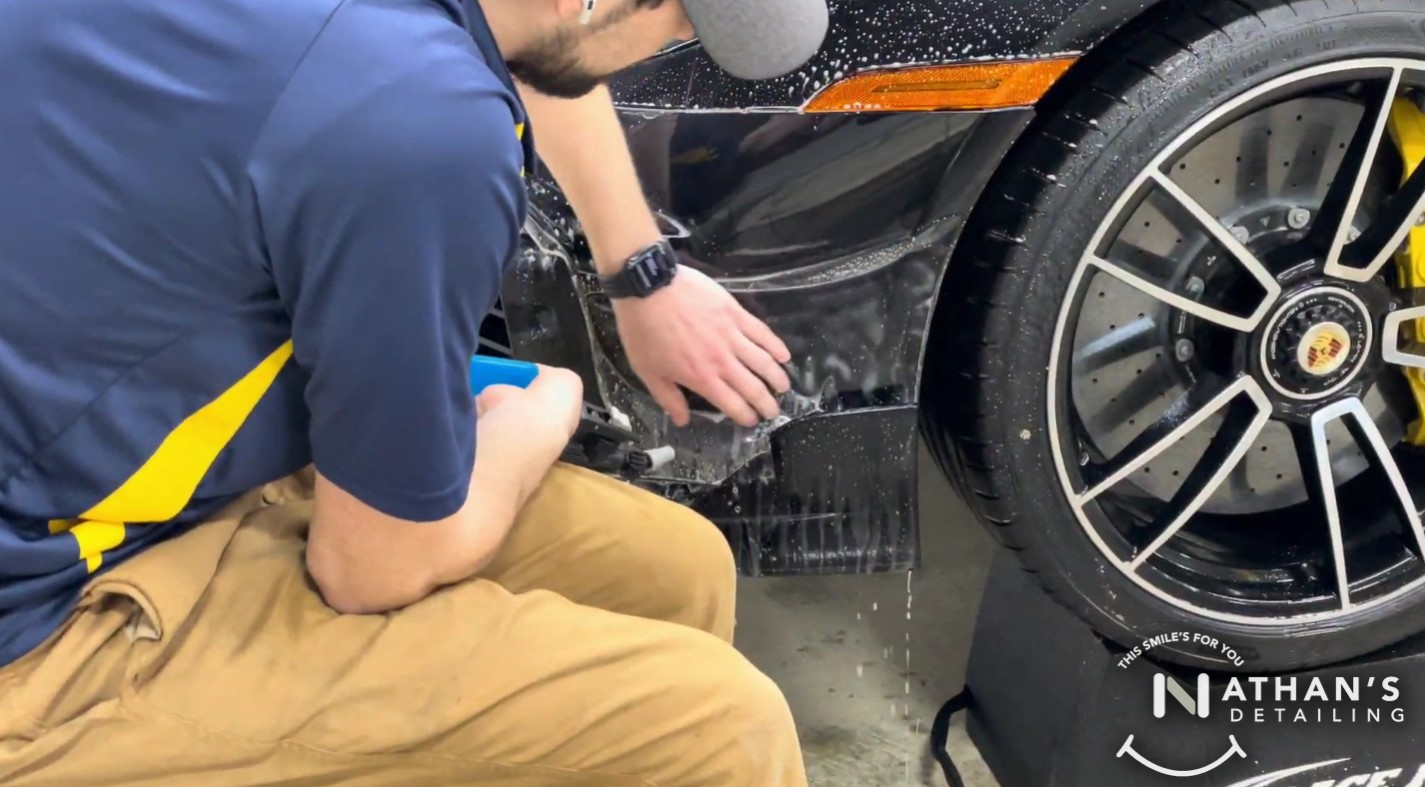 Technician applying paint protection film to the lower side panel near a Porsche wheel.