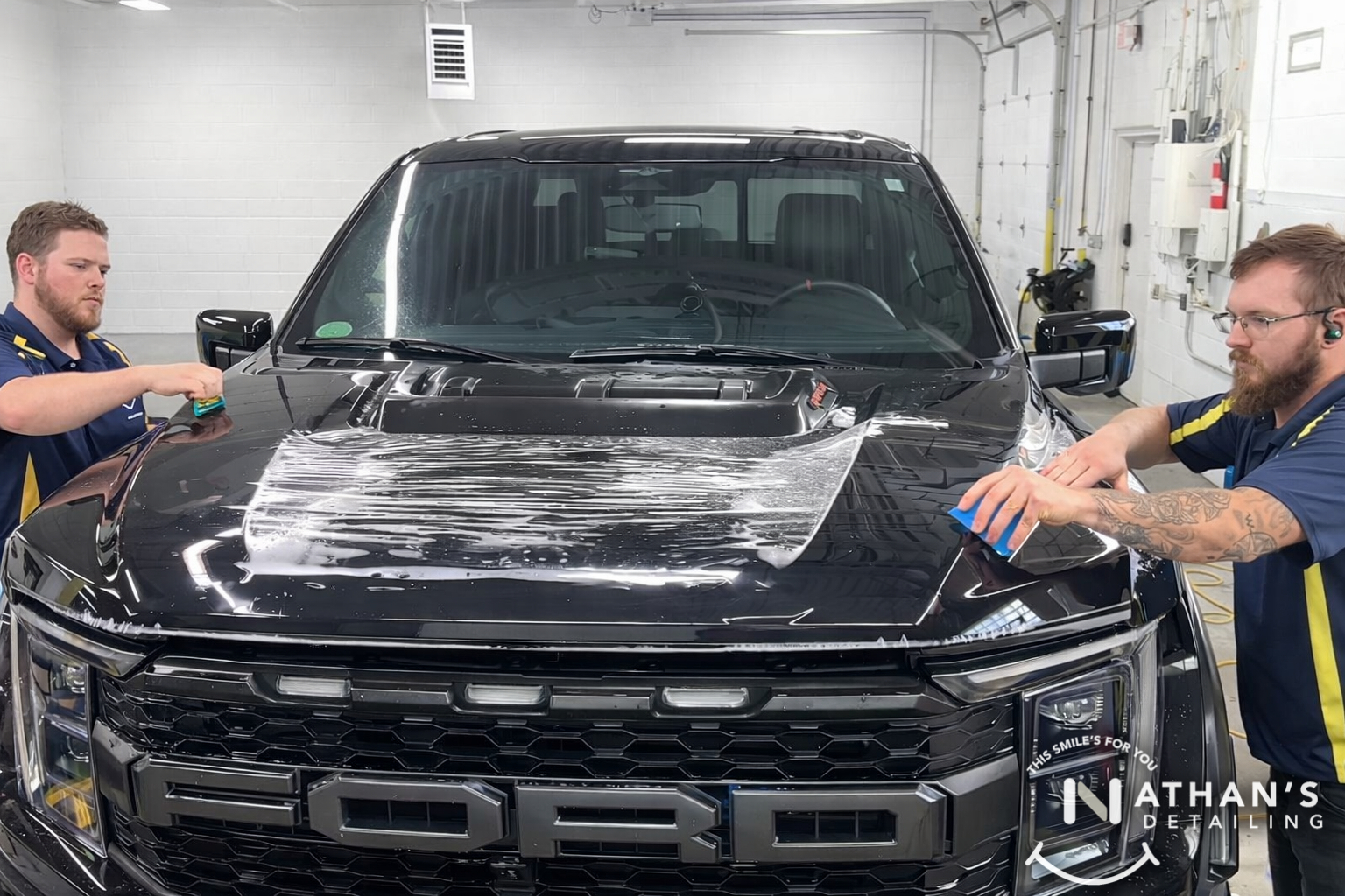 Two technicians applying paint protection film across a black truck hood.