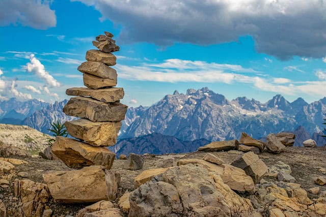 A carefully balanced stack of stones on a rocky hilltop, with a panoramic view of rugged mountain ranges under a vibrant blue sky dotted with clouds.