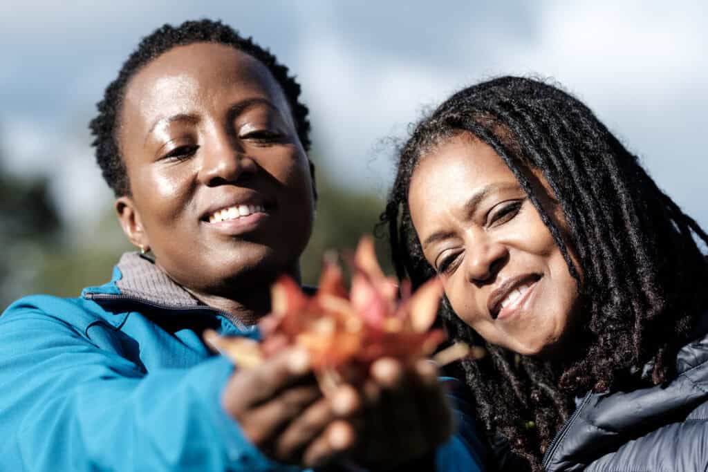 Two smiling women stand closely outdoors, joyfully holding a handful of blurred autumn leaves toward the camera. The sky is partly cloudy behind them, and they appear to be enjoying a moment of connection with nature.