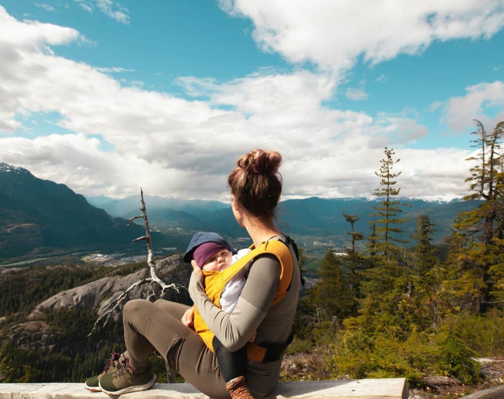 A serene outdoor moment of a woman sitting on a log in a mountainous landscape, holding a baby in a carrier. The setting is with lush green forests, towering mountains, and a bright blue sky with scattered clouds. The woman is looking out at the expansive view and the baby, snugly secured, gazes outward.