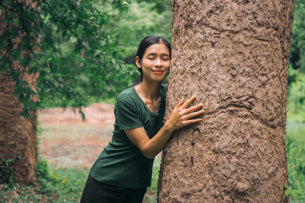 A young woman embraces a large tree with her eyes closed and a peaceful smile, surrounded by lush green foliage, expressing a deep sense of connection with the natural environment.
