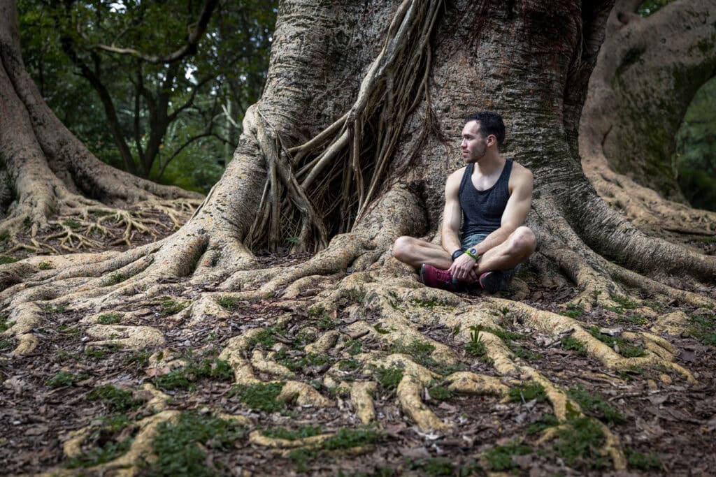A man in athletic wear sits cross-legged at the base of a massive tree with sprawling roots in a forest, gazing thoughtfully into the distance, suggesting mindfulness and grounding.