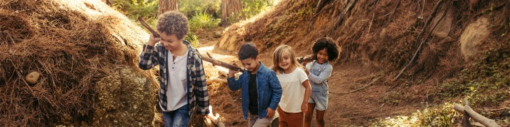 Four young children carrying sticks while walking through a forested area, emphasising teamwork and adventure.
