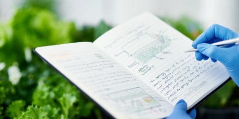 Scientist wearing blue gloves writing research notes in a notebook with plant illustrations, surrounded by green leafy plants in a greenhouse.