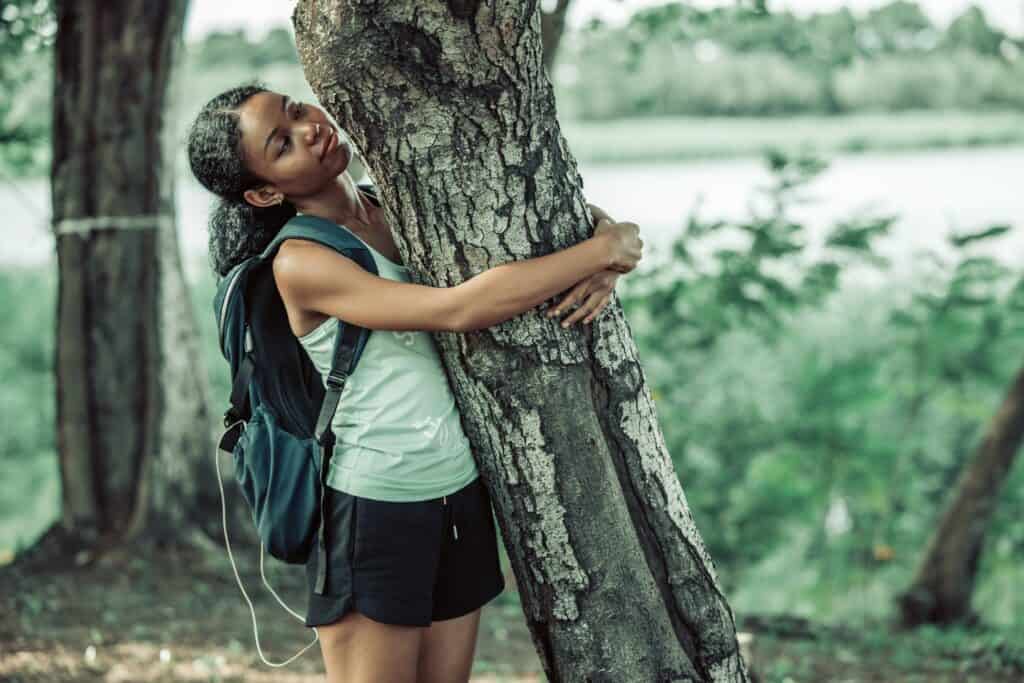 A young woman with a backpack is embracing a tree in a peaceful, forested area, her eyes closed as she connects with nature.