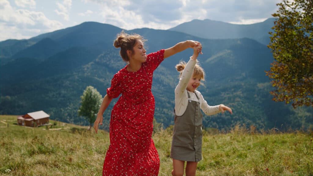A young woman in a red dress dances joyfully with a younger girl in the mountains, surrounded by green hills and blue peaks.