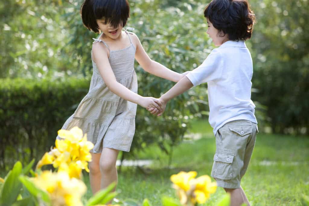 Two young children joyfully spin in a circle holding hands in a green garden filled with yellow flowers.