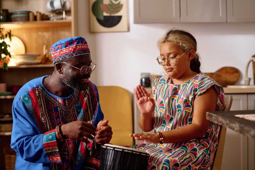 An elderly man in traditional attire and a young girl wearing a colourful dress are sitting together, playing a drum. The man is smiling while teaching the girl how to play, creating a warm, intergenerational moment.