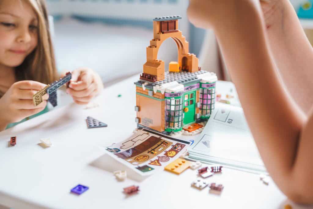 Two young children assembling a detailed LEGO building set at a white table, surrounded by small pieces, instruction booklets, and focused expressions.