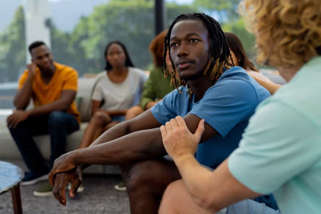 A young man sits attentively in a group circle, listening with a thoughtful expression as another person places a reassuring hand on his arm. Others in the background appear engaged in the supportive and reflective atmosphere.