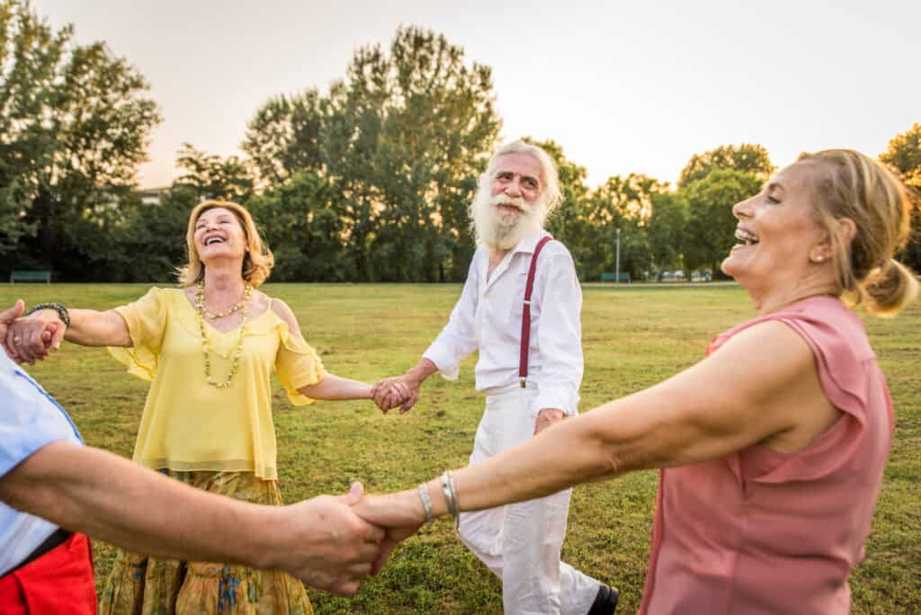 A group of seniors holds hands in a circle, laughing and dancing together on a grassy field at sunset. Their joyful expressions and flowing movements capture a moment of community, vitality, and connection in nature.