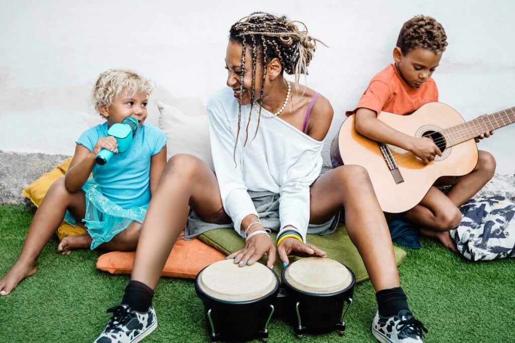 A joyful family jam session with a woman playing bongos, a child singing into a toy microphone, and another playing guitar on the grass.