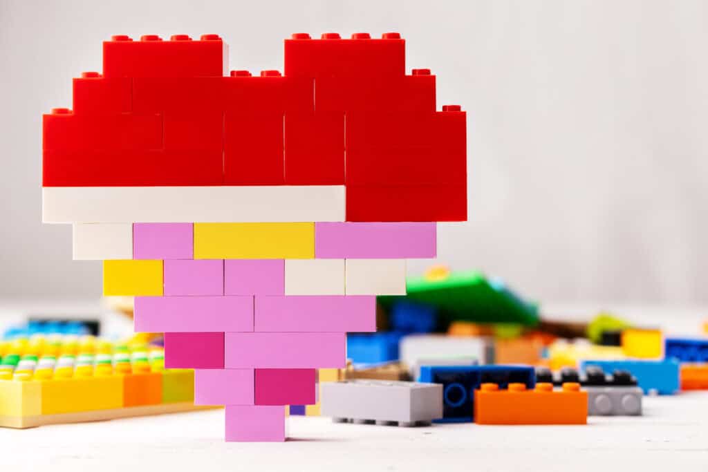 Heart-shaped structure built from red, pink, yellow, and white plastic bricks, standing upright on a table with other colourful building blocks scattered in the background.