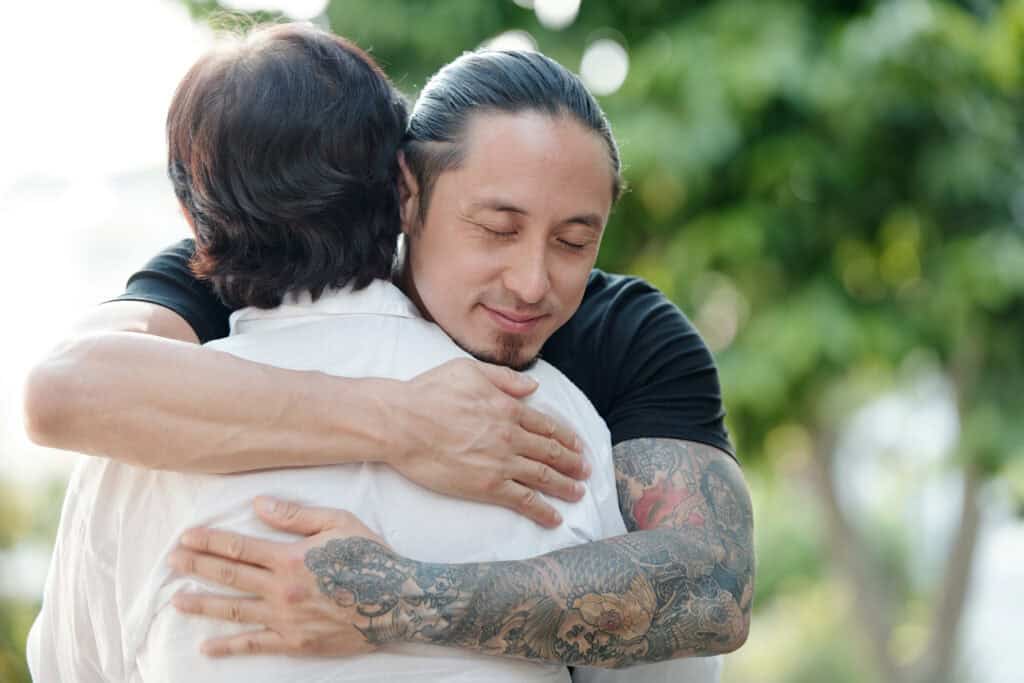 A man with tattoos and a black shirt embraces another person in a white shirt, eyes closed and smiling softly, expressing warmth, trust, and emotional connection in a natural outdoor setting.