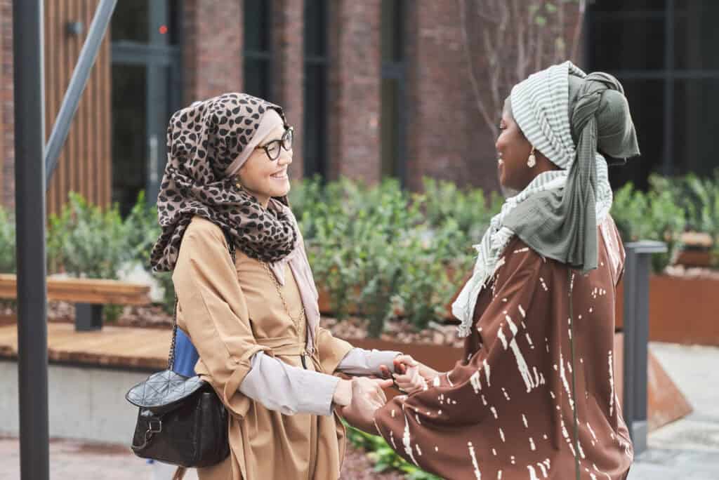 Two women wearing hijabs warmly greet each other outdoors, smiling and holding hands. They stand in a courtyard area with greenery and modern architecture in the background, radiating connection, respect, and friendship.
