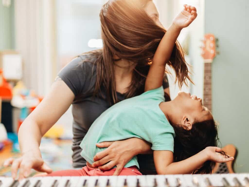 A mother and her daughter are playfully enjoying music together at home. The mother is holding her daughter while playing a piano, and the child is joyfully leaning back, laughing.