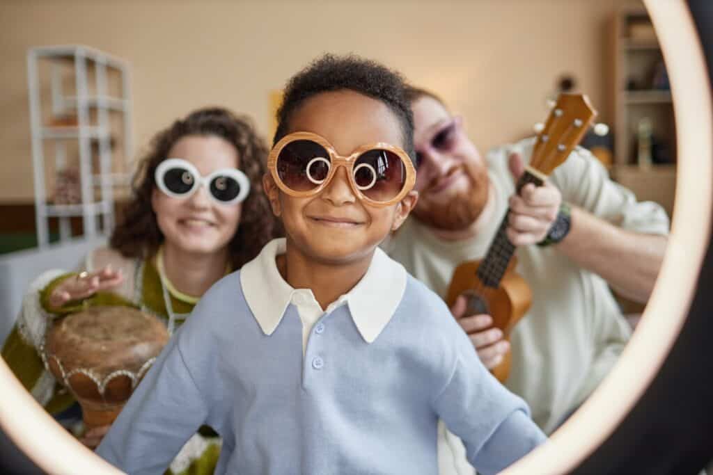 A young boy with oversized sunglasses is smiling in front of a ring light, with two adults behind him holding musical instruments, including a drum and a ukulele, adding a playful, family-oriented vibe.