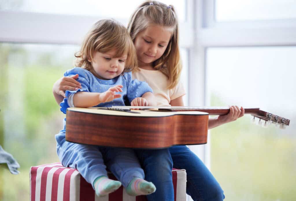 An older girl lovingly guides her toddler sibling’s hand on a guitar as they sit together by a window.