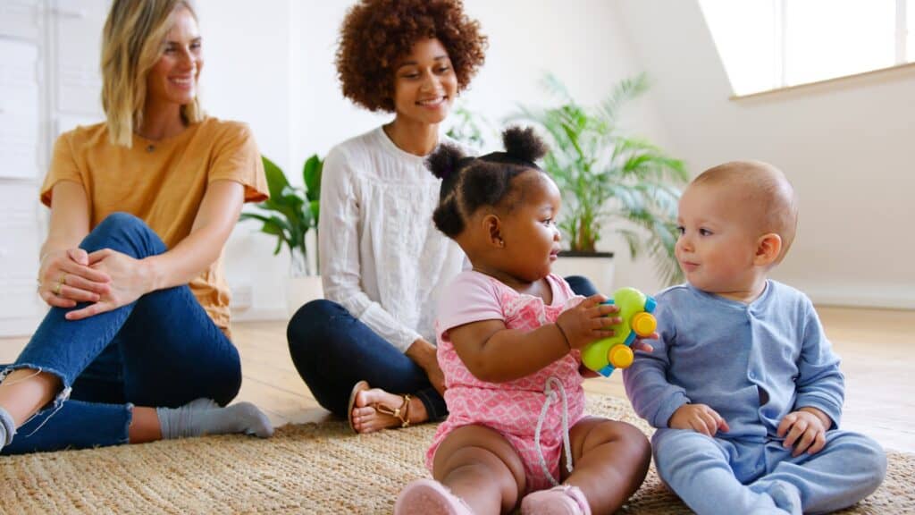 Two mothers sit on a rug in a bright, modern room, smiling as they watch their babies interact. One baby, wearing pink, holds a colorful toy while facing another baby dressed in light blue.