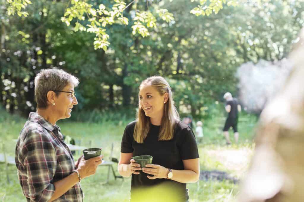 Two women standing in a sunlit meadow, holding cups and engaged in friendly conversation, while children play in the background.