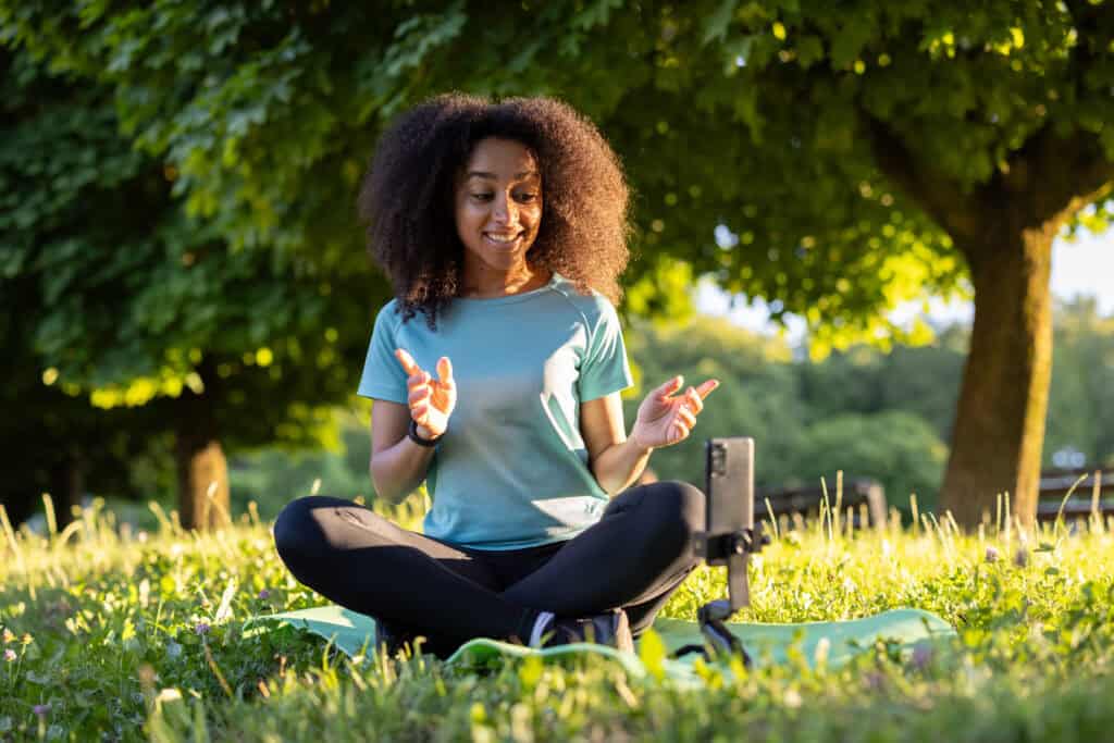 A smiling woman in a teal sports top and leggings sits on a yoga mat in a park, recording a video with her phone on a tripod.