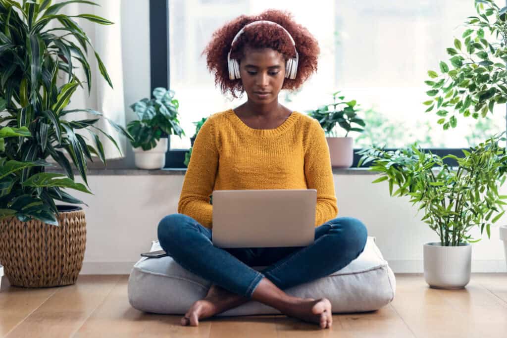 A young woman in a mustard sweater and jeans sits cross-legged on a cushion, working on her laptop while wearing headphones, surrounded by green plants.
