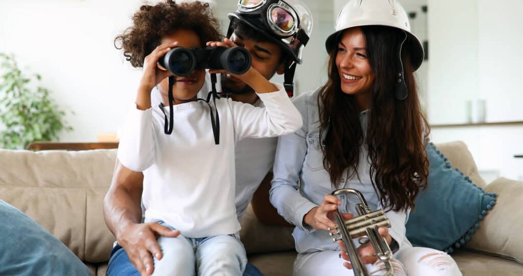 A happy family engaged in imaginative play, with a young girl using binoculars while her parents, dressed in fun hats, smile beside her.