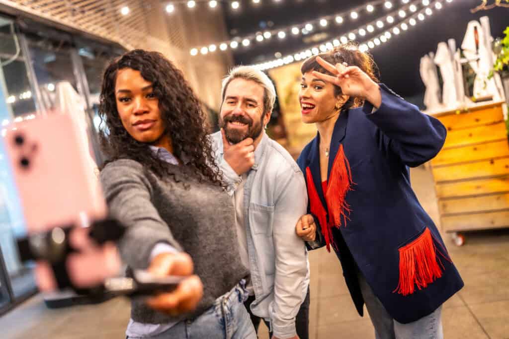 Three friends taking a selfie together in an outdoor setting with string lights, smiling and posing playfully.