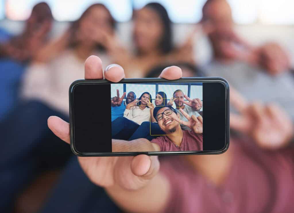 A hand holding a smartphone showing a group selfie, with the actual group blurred in the background, all making peace signs.