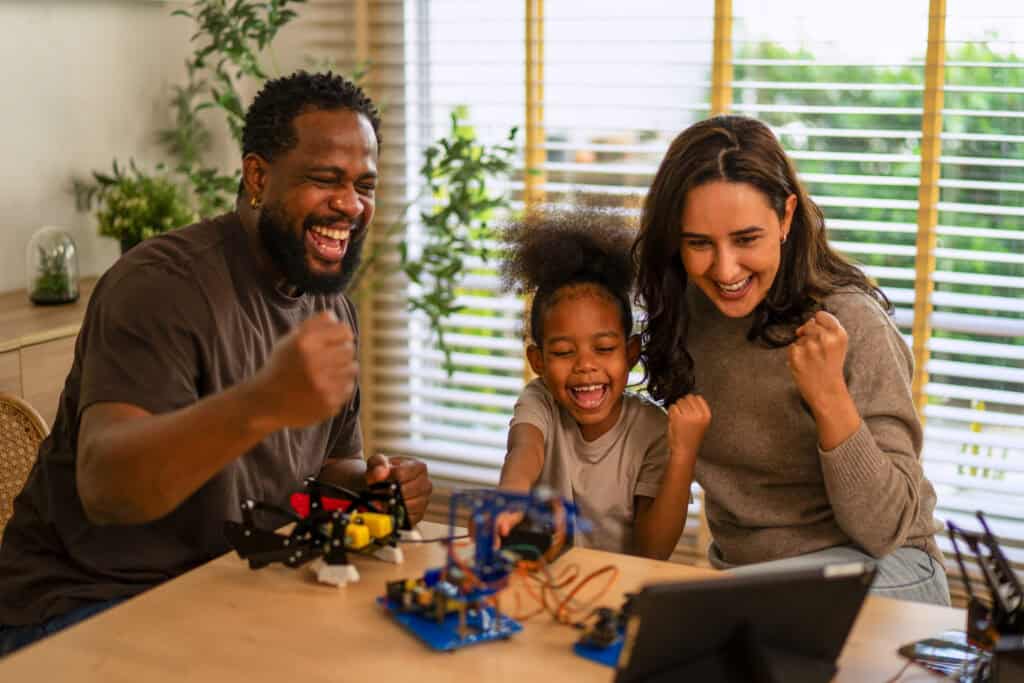 A joyful familyโfather, mother, and young daughterโcelebrating together while working on a robotics project at home, with a tablet on the table.