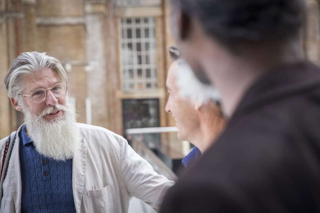 An older man with a long white beard, glasses, and a beige jacket talks to two other men at a train station, smiling and gesturing warmly.