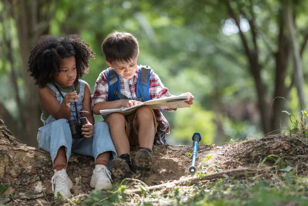 Two children sit under a tree, one holding binoculars and the other reading a map, surrounded by nature.