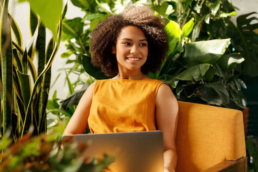 A young woman with curly hair wearing an orange dress sits on an orange chair, smiling while using her laptop, surrounded by lush green plants.