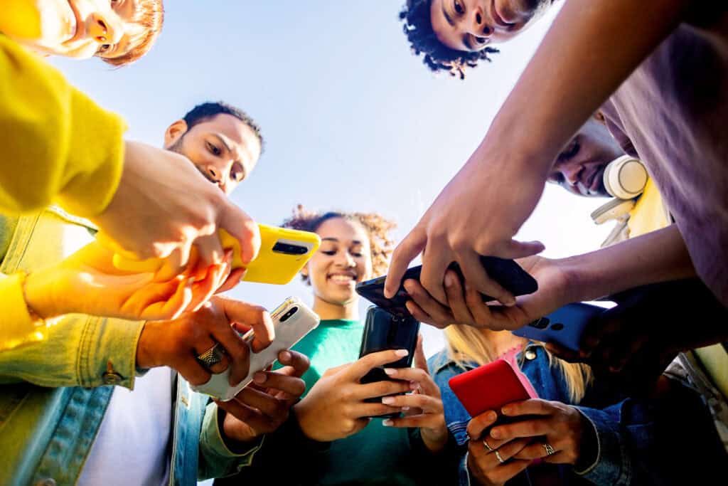 A diverse group of young people standing in a circle, looking at their smartphones, smiling and engaged.