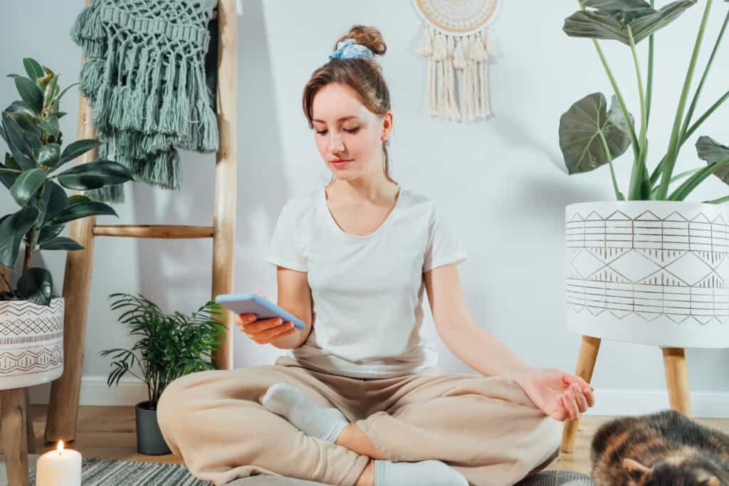 A woman in a white shirt and beige pants sits in a meditative pose, holding a phone, in a cosy room with plants, candles, and macrame decor.