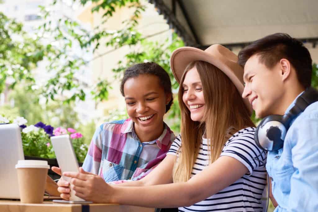 Three diverse young adults sitting at an outdoor cafรฉ, laughing and looking at a tablet together.