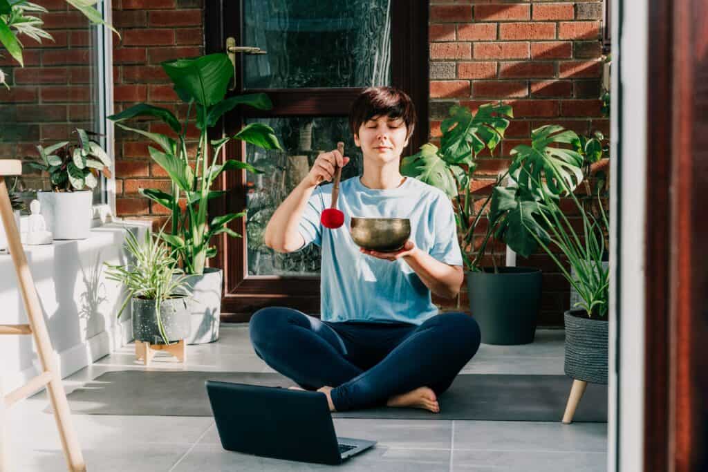 A woman in a blue shirt and leggings sits on a yoga mat in a sunlit indoor space, using a Tibetan singing bowl for meditation.