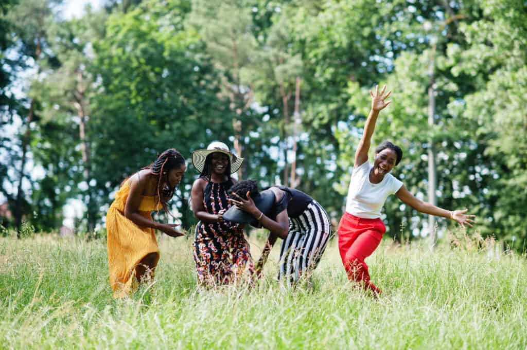 Four women in vibrant summer clothing laugh and dance in a grassy meadow surrounded by trees, radiating joy and playful movement.