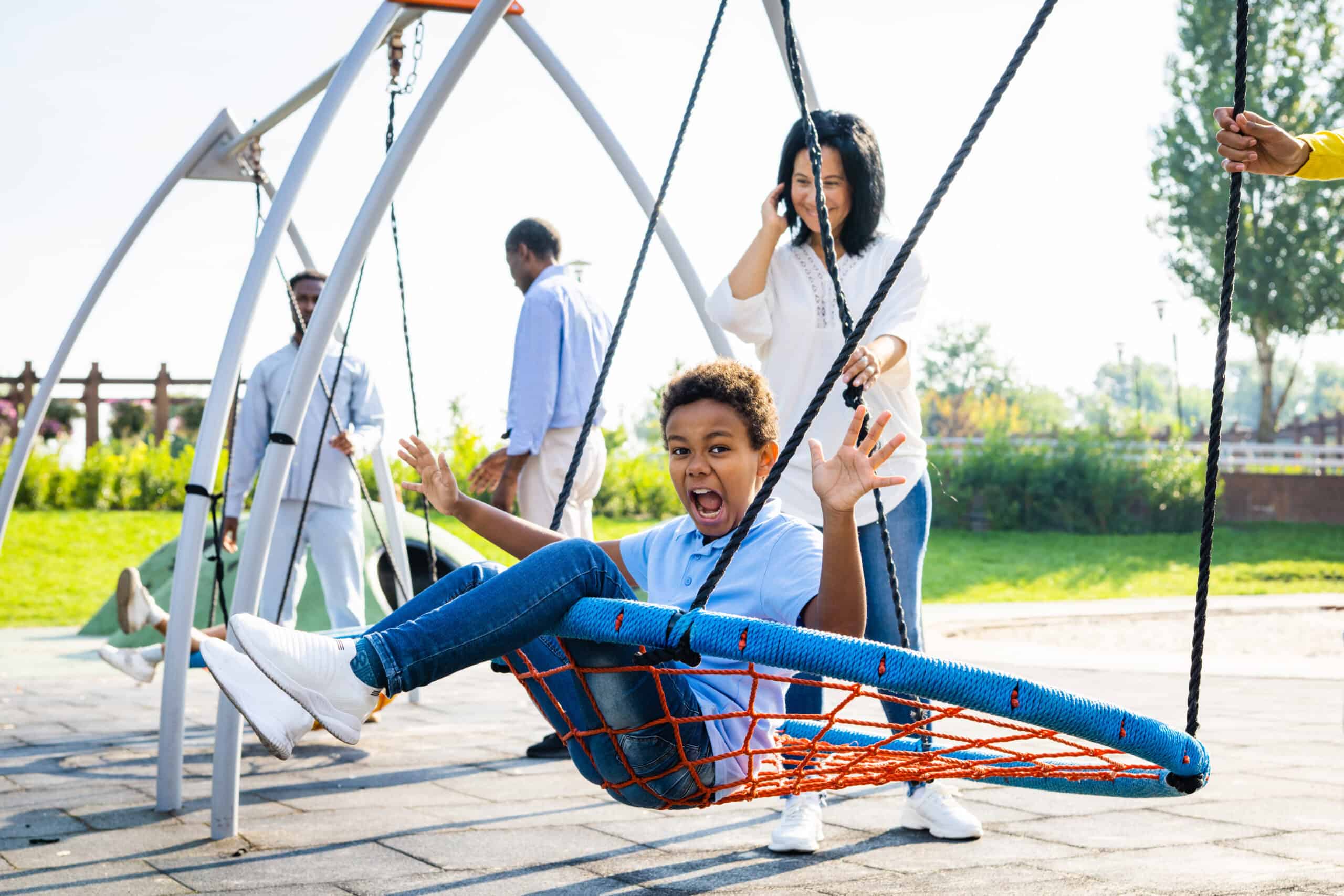 A young boy gleefully swings on a large rope net swing at a playground, pushed by an adult while others stand nearby in the sunshine.