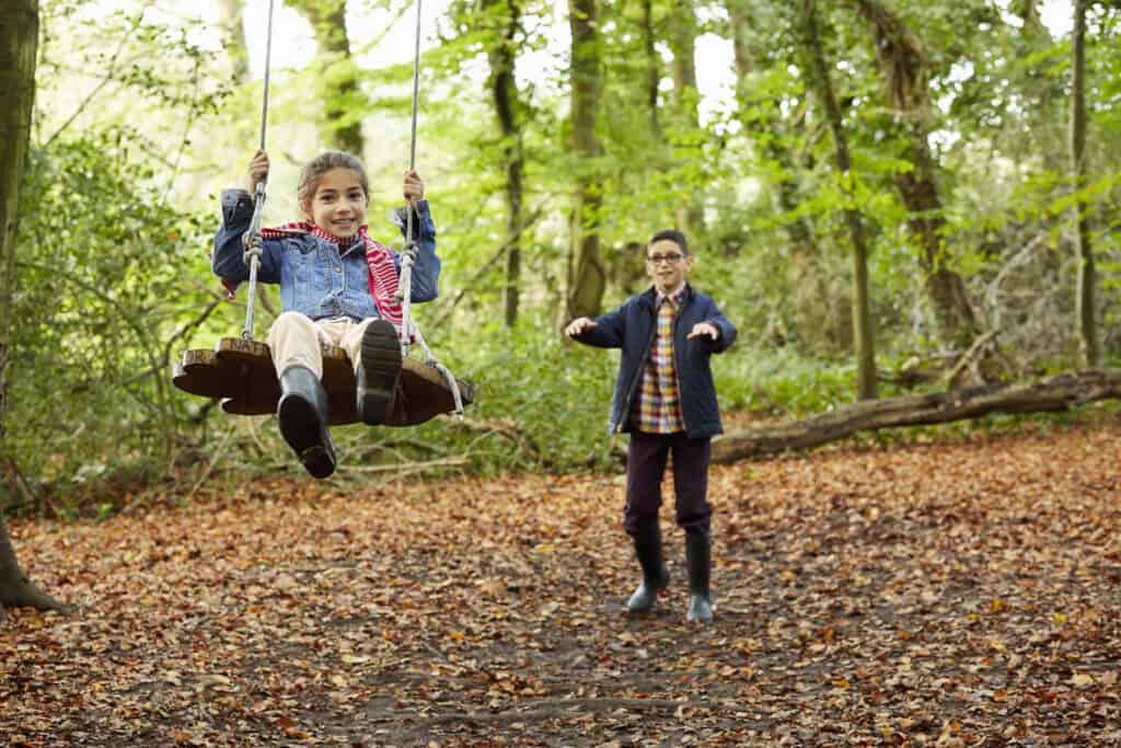 A young girl smiles while sitting on a wooden swing suspended from tree branches in an autumn forest. A boy stands behind her, gently pushing the swing. The ground is covered with fallen leaves, and the background is filled with tall trees and green foliage, capturing a joyful moment of outdoor play and sibling connection.