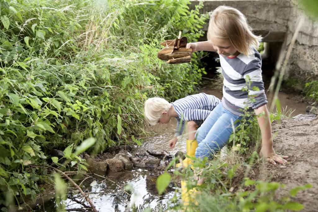 Two young children explore a small creek, one holding a handmade wooden boat and the other crouching to play in the water, surrounded by lush greenery.